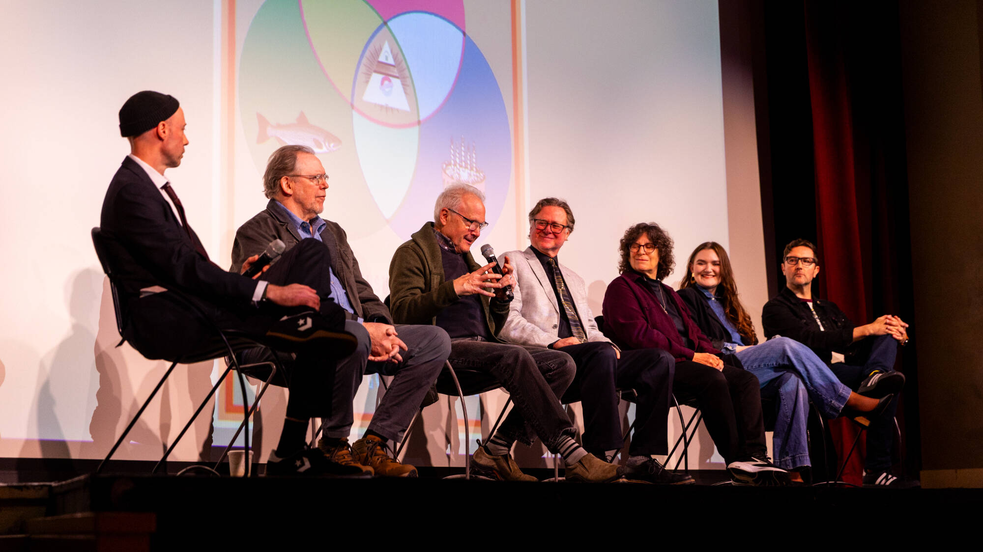 Six individuals sit on a stage in front of a colorful projected graphic, engaging in a panel discussion.